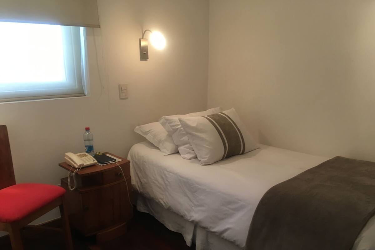 A hotel room with a bed, a bedside table with a phone, a chair, and a window.