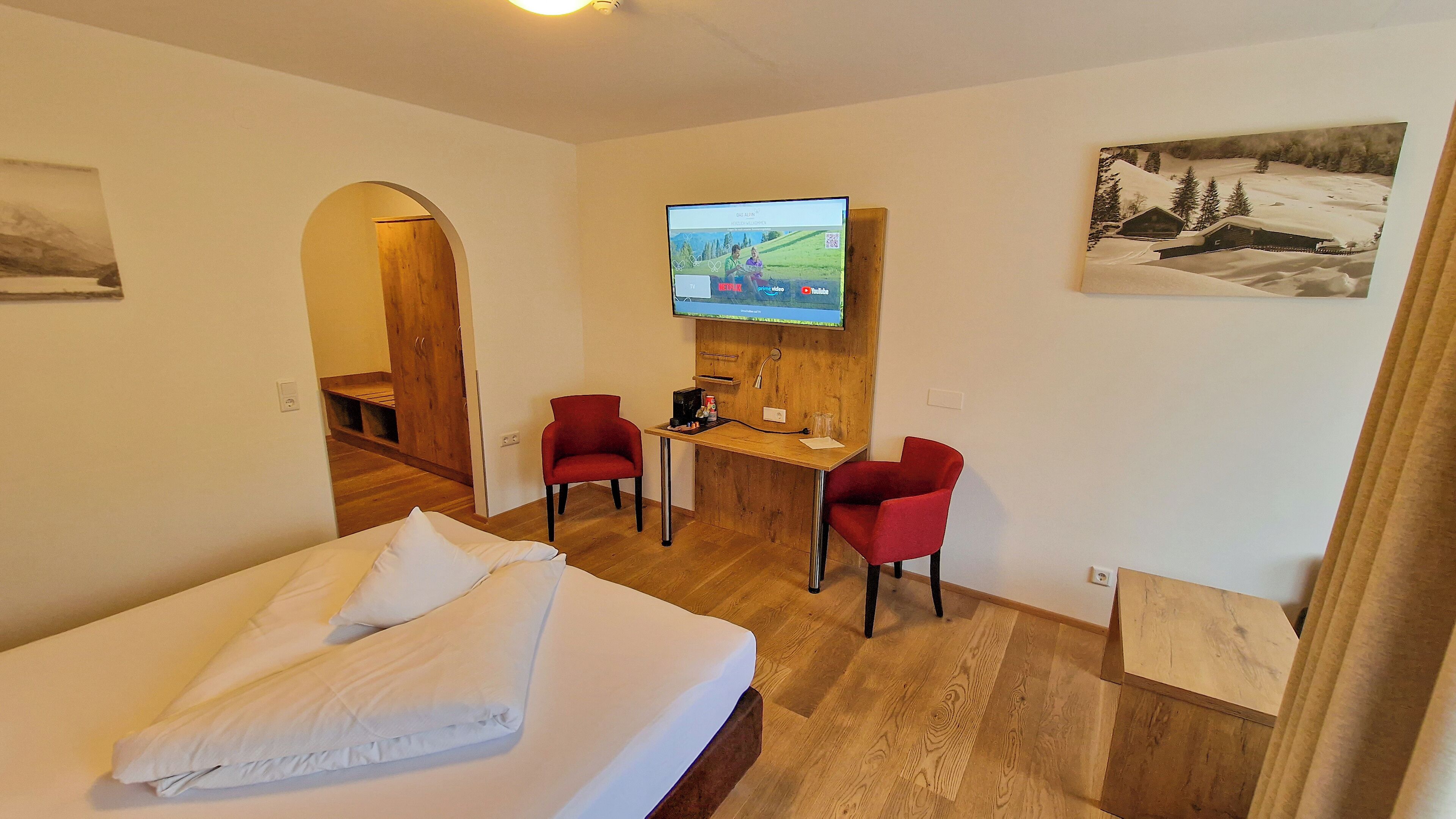 Een hotelkamer met een bed, twee rode stoelen, een bureau met een computer, een televisie en een schilderij aan de muur.