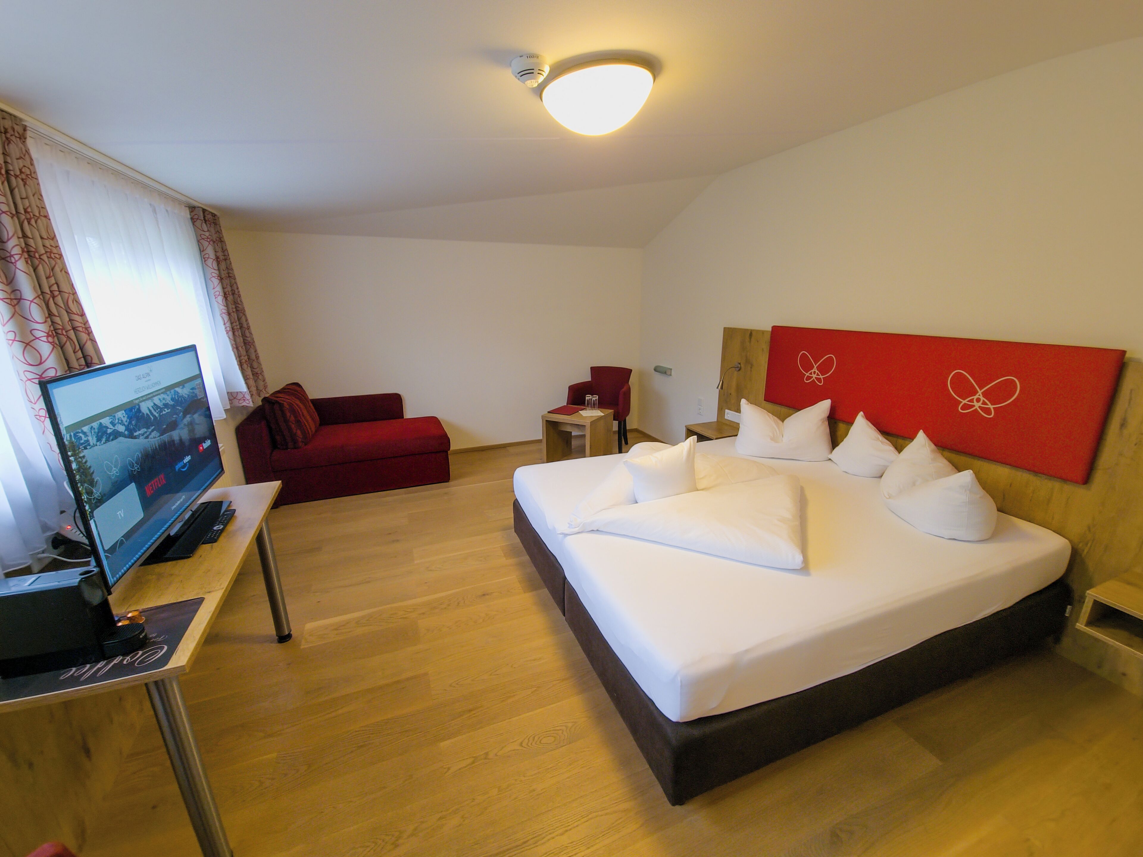Une chambre d’hôtel avec un lit, un bureau avec un ordinateur, une télévision et un fauteuil rouge.