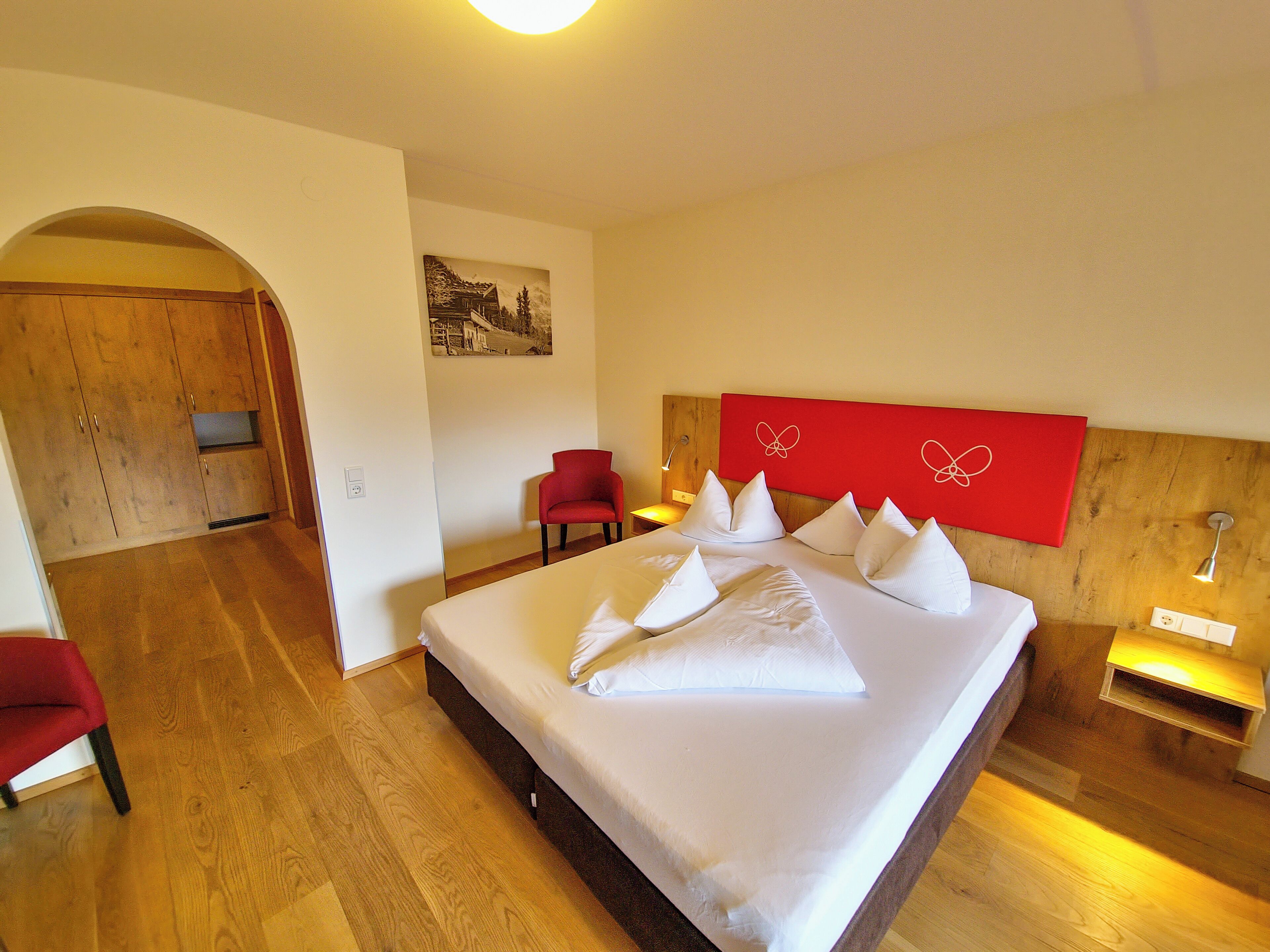 Habitación de hotel con una cama grande, una silla roja, un armario de madera y un cuadro en la pared.