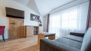 Ein modernes Wohnzimmer mit Holzboden, einem grauen Sofa, einer Holzkommode, einem Flachbildfernseher und einem Fenster mit durchsichtigen Vorhängen.
