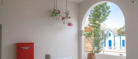 A balcony with a white railing, a red cabinet, a white bench, and a view of blue buildings and a tree through an archway.