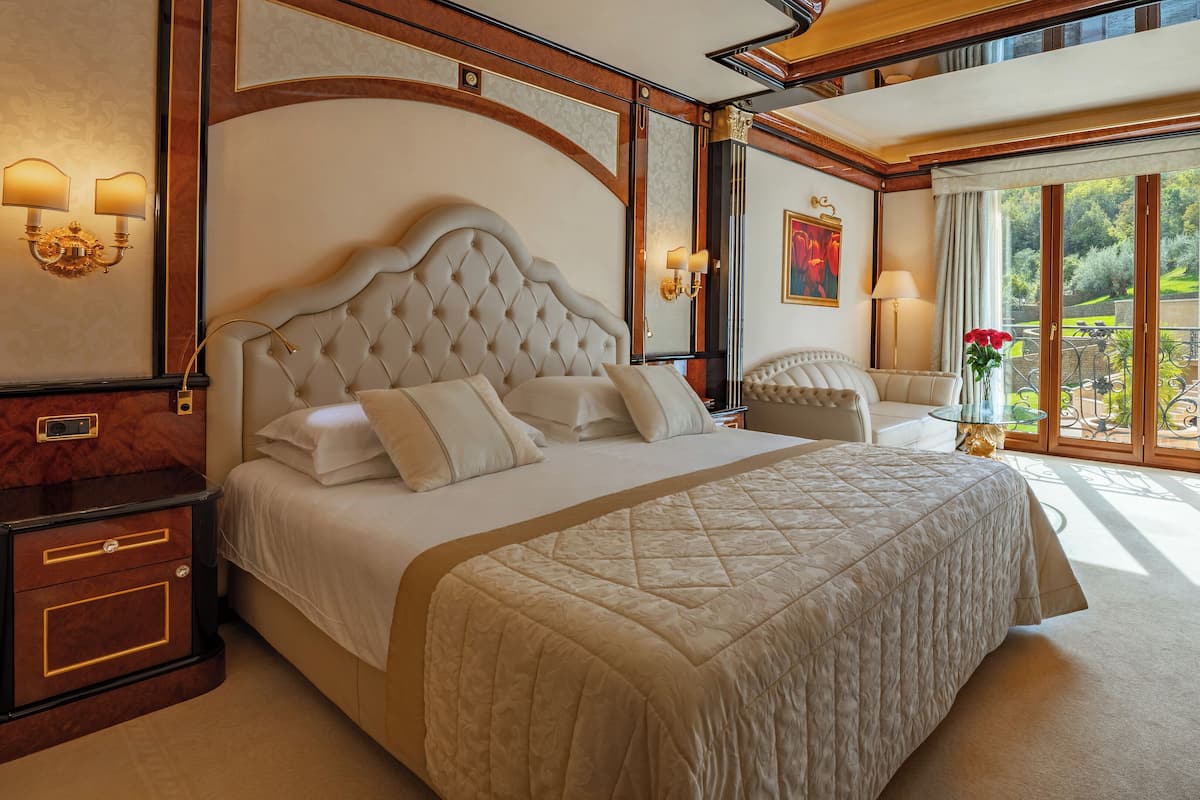 A luxurious bedroom with a large bed, a chaise lounge, a glass table, and a view of a garden through a sliding glass door.