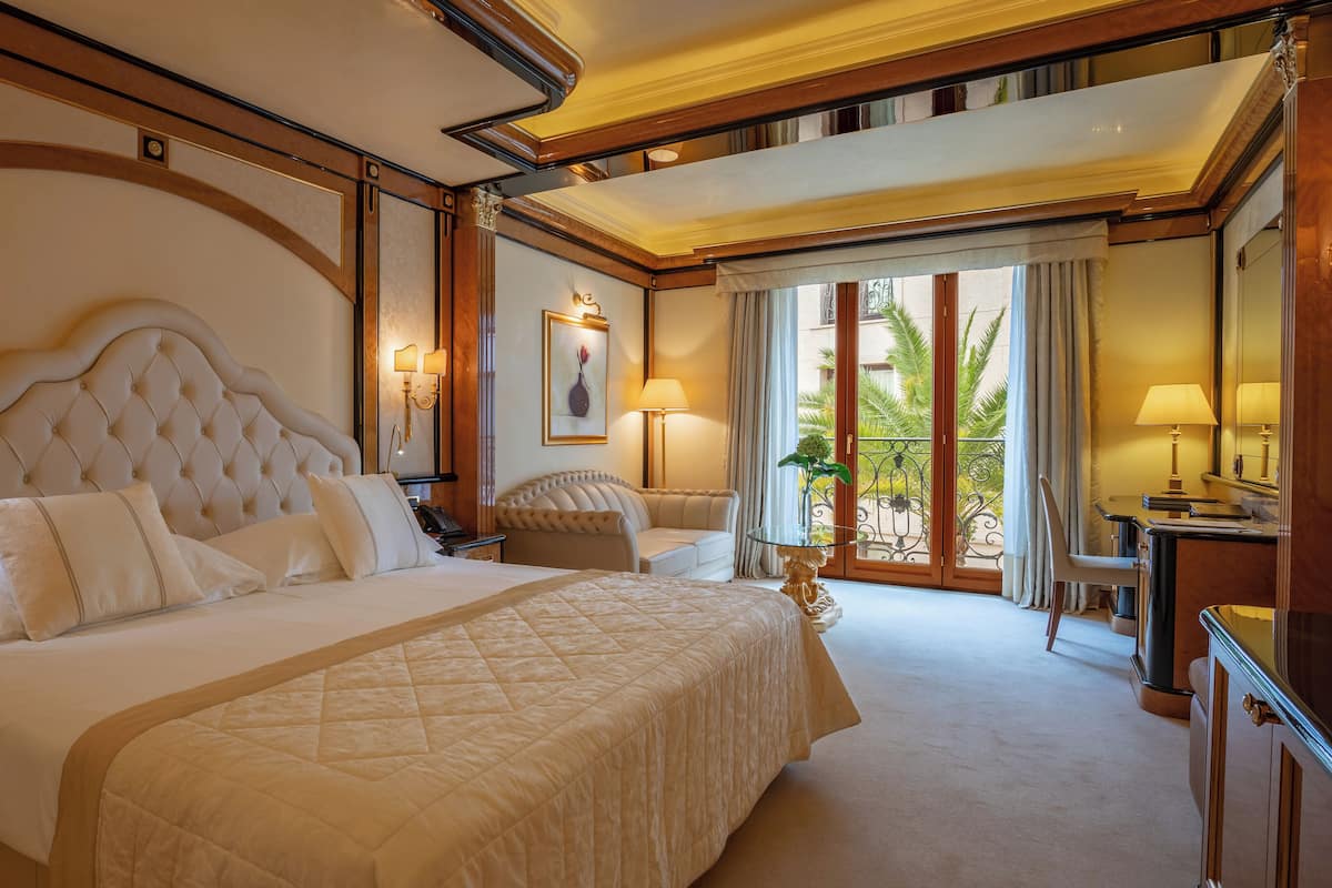 A luxurious hotel room with a large bed, a sitting area with a chair and table, a desk with a lamp, and a balcony with a view of palm trees.