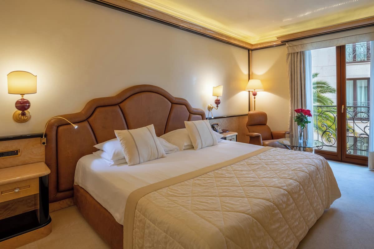 A hotel room with a large bed, two bedside tables, a chair, and a view of a balcony with a railing.