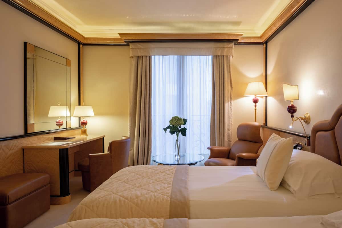 A hotel room with a large bed, a desk, a chair, a small table with a vase, and a window with curtains.