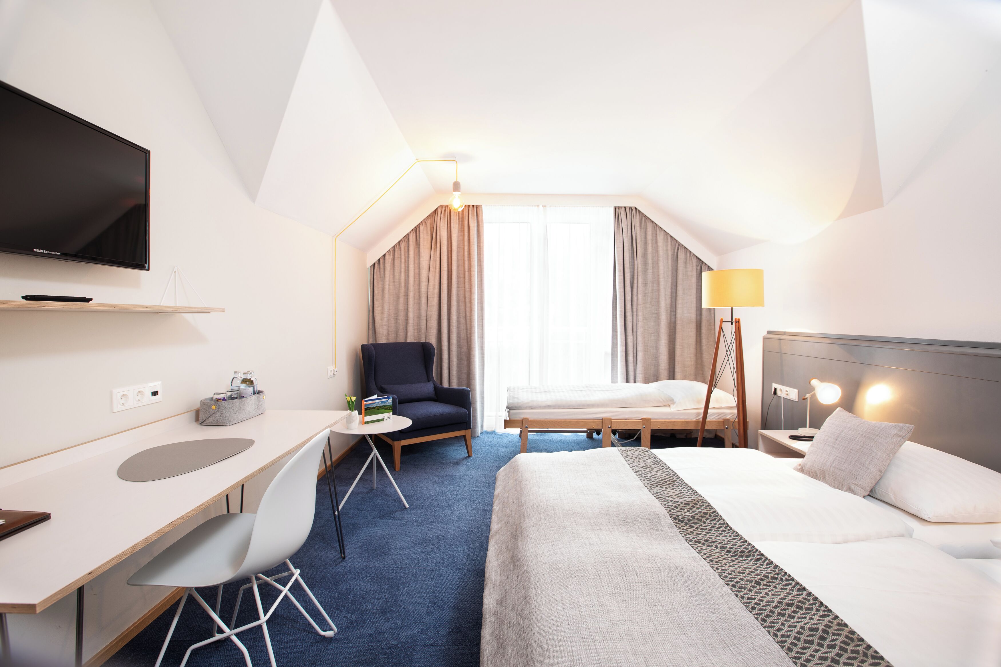 A hotel room with a bed, desk, chair, TV, and a lamp.