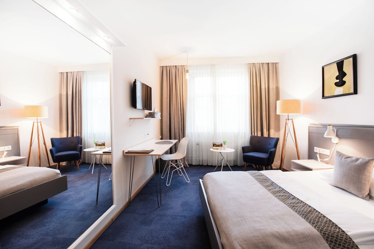 A hotel room with a large bed, a desk, two chairs, a TV, and a small table with a lamp.