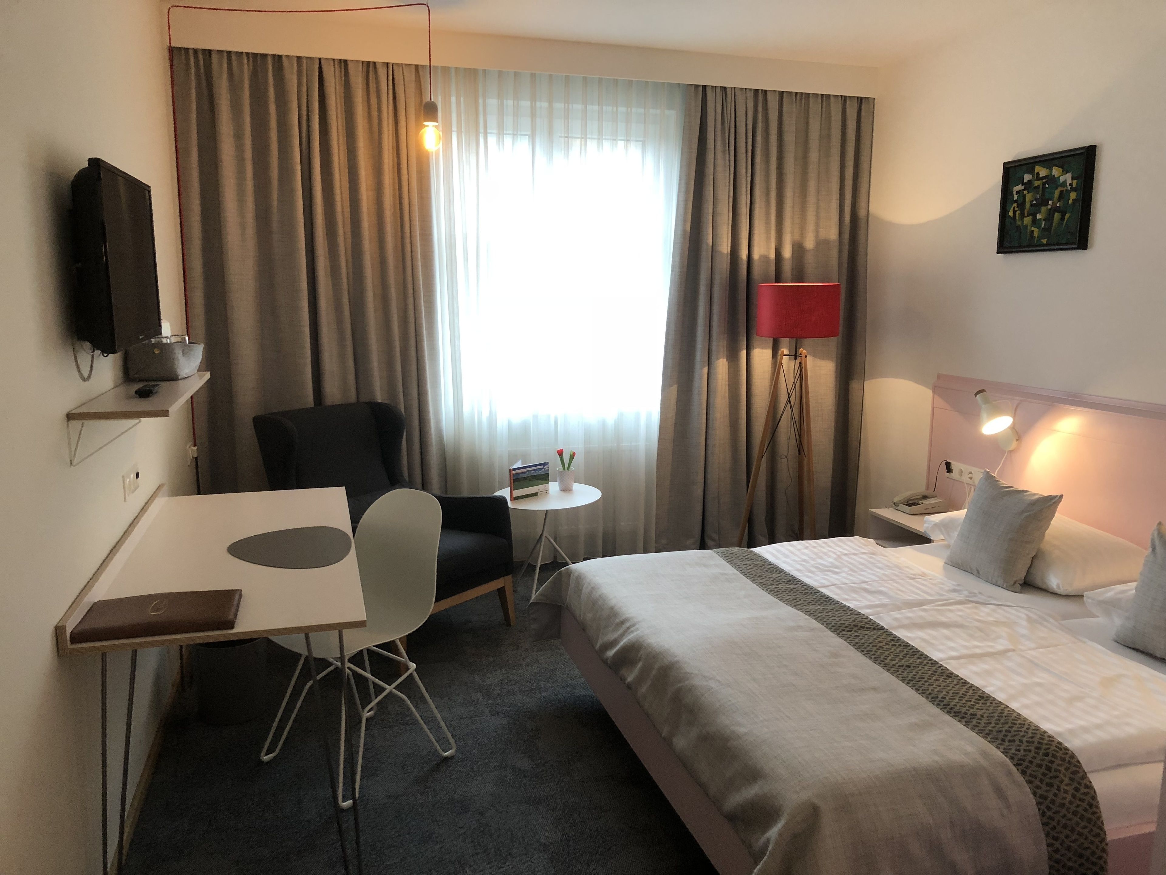 Ein Hotelzimmer mit einem Bett, einem Schreibtisch, einem Stuhl, einem kleinen Tisch, einer Lampe und einem Bild an der Wand.