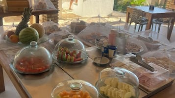 Buffet com diversos alimentos cobertos, incluindo frutas, doces e carnes, sob uma pérgola de madeira, com vista para um jardim e área de estar ao ar livre.