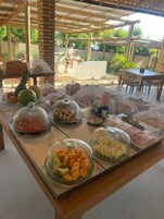 Buffet com diversos alimentos cobertos, incluindo frutas, doces e carnes, sob uma pérgola de madeira, com vista para um jardim e área de estar ao ar livre.
