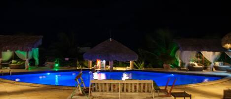 A nighttime swimming pool area with a thatched-roof structure, lounge chairs, and a well-lit pool.