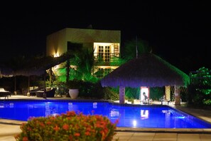 Una vista notturna di un resort con piscina, costruzioni a tetto di paglia e un edificio ben illuminato.