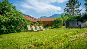 Uma área externa gramada com espreguiçadeiras e um prédio de madeira com telhado vermelho, cercado por árvores e montanhas ao fundo.