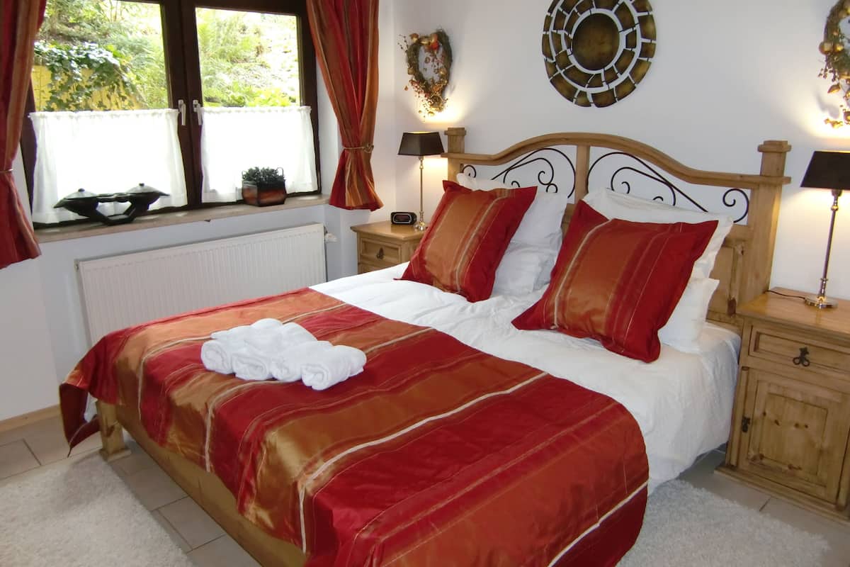 Una cama bien hecha con colcha roja y blanca, una toalla blanca y dos lámparas de noche sobre mesitas de madera.