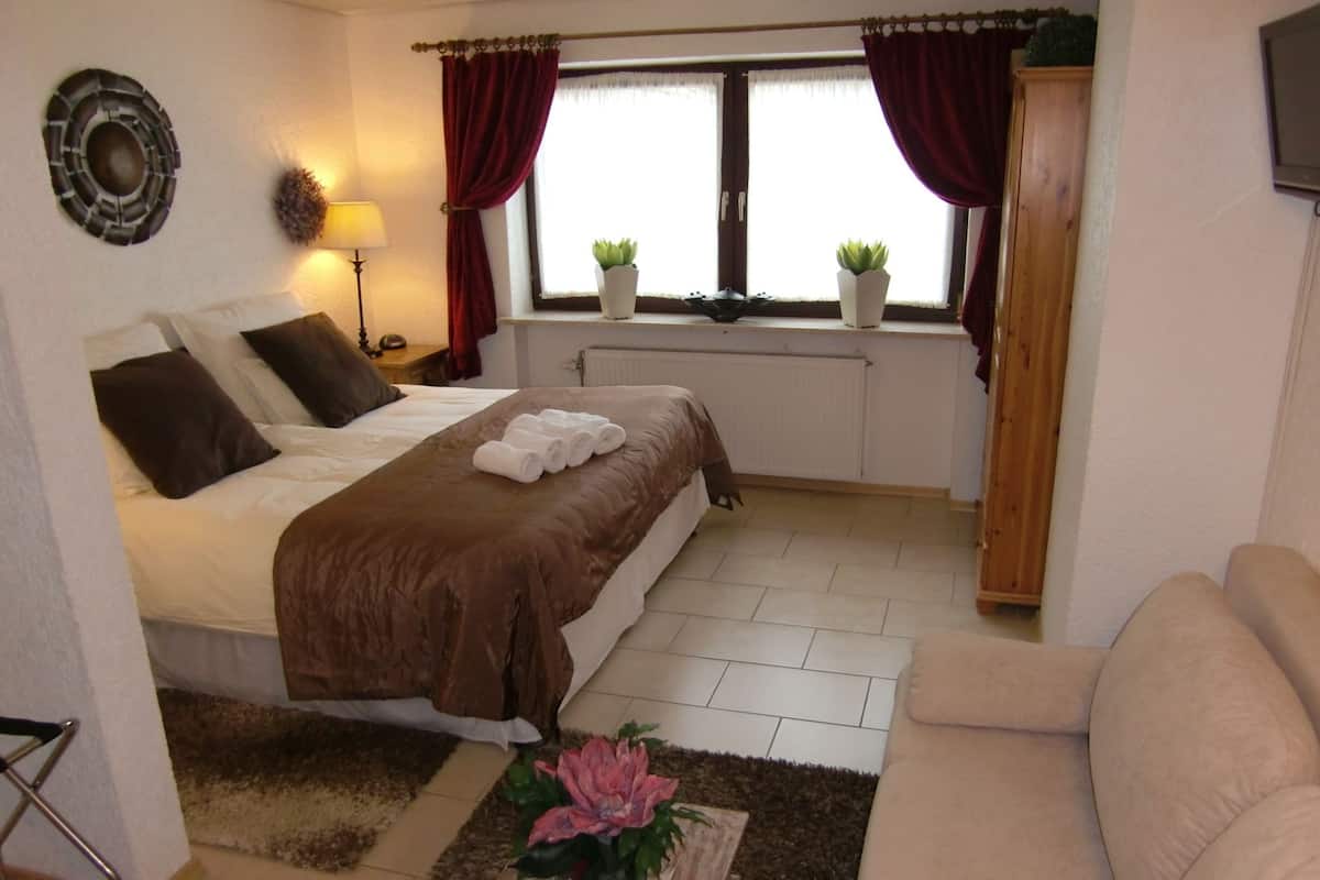 Un dormitorio bien iluminado con una cama, una mesita de noche con lámpara, una ventana con cortinas, un radiador, un armario de madera y un sofá.