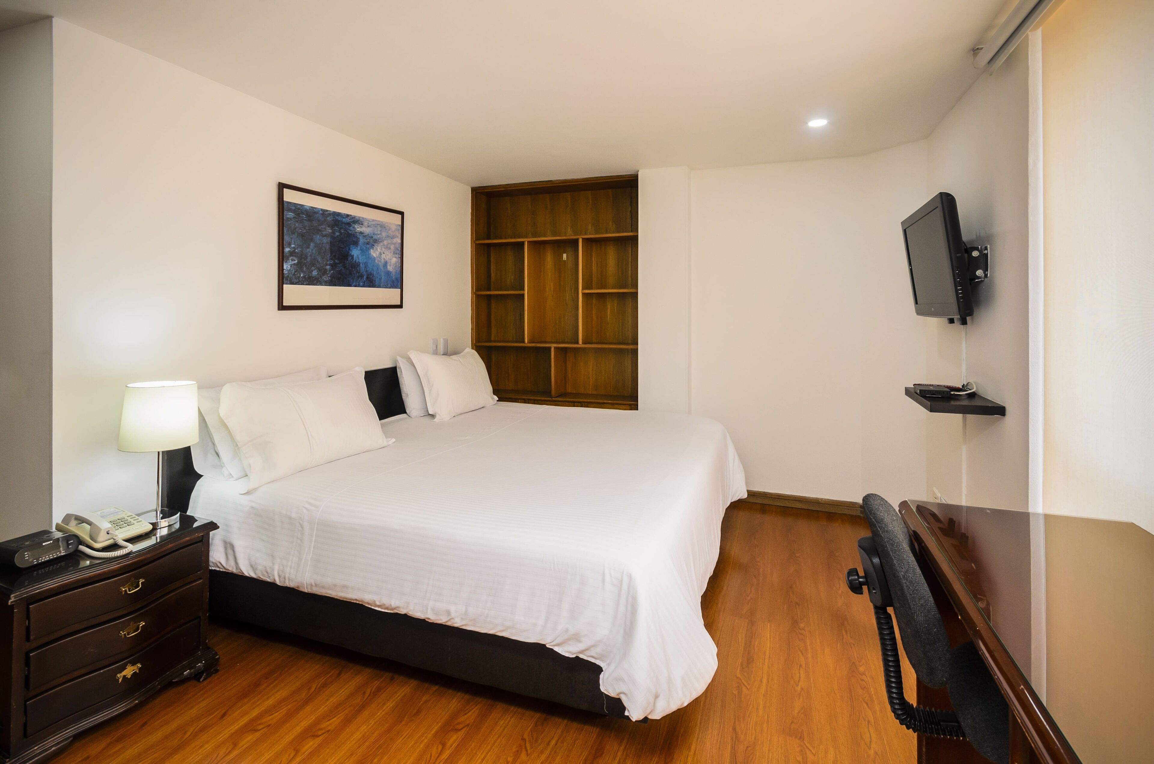 A neatly arranged hotel room with a large bed, bedside table with a lamp, a wooden shelf unit, and a mounted television.