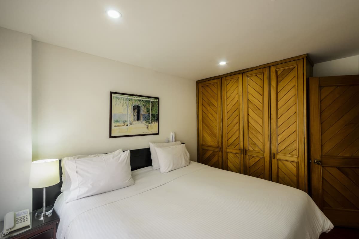 Ein Hotelzimmer mit Bett, Nachttisch, Lampe, einem Gemälde an der Wand und Holztüren zu einem Kleiderschrank.