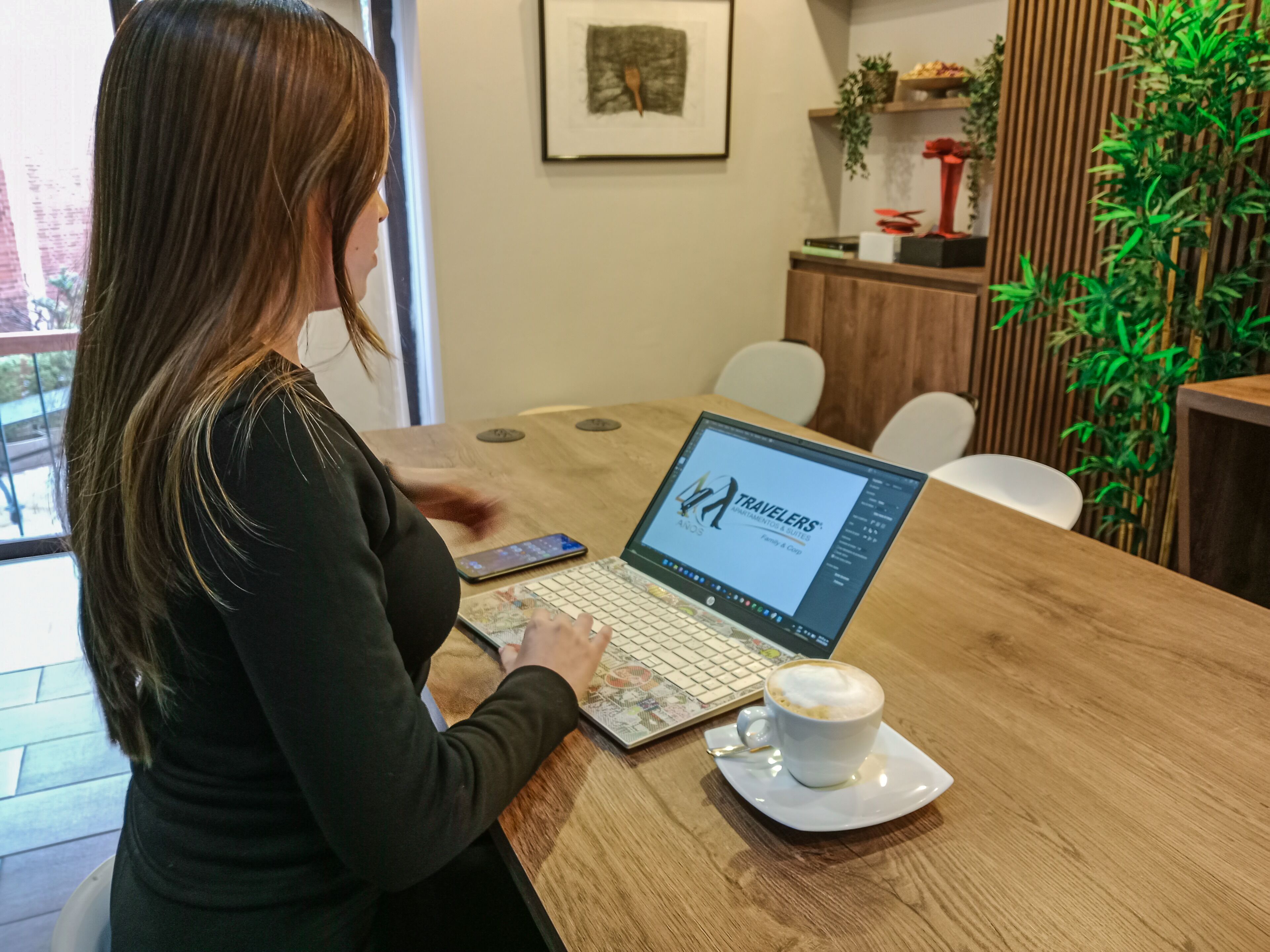 A person using a laptop at a wooden table with a cup of coffee, in a room with a plant and a framed picture on the wall.