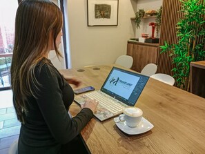 Eine Person benutzt einen Laptop an einem Holztisch, neben einer Kaffeetasse, in einem Raum mit einer Pflanze und einem gerahmten Bild an der Wand.