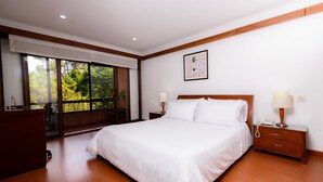 A bedroom with a large bed, wooden headboard, bedside tables with lamps, a desk, and a window with a view of trees. - Travelers Suites Condominio Plenitud  (Bogotá)