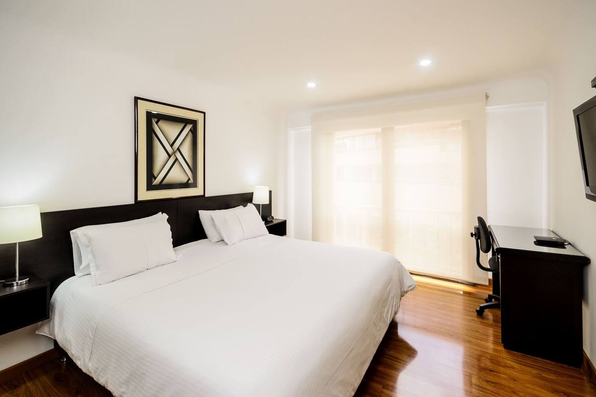 A neatly made bed with white linens, a dark headboard, and a framed artwork above it.