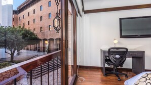 A room with a desk, chair, and a TV on the wall. There is a balcony with a railing and a view of a building with multiple windows. - Travelers Suites Condominio Plenitud  (Bogotá)