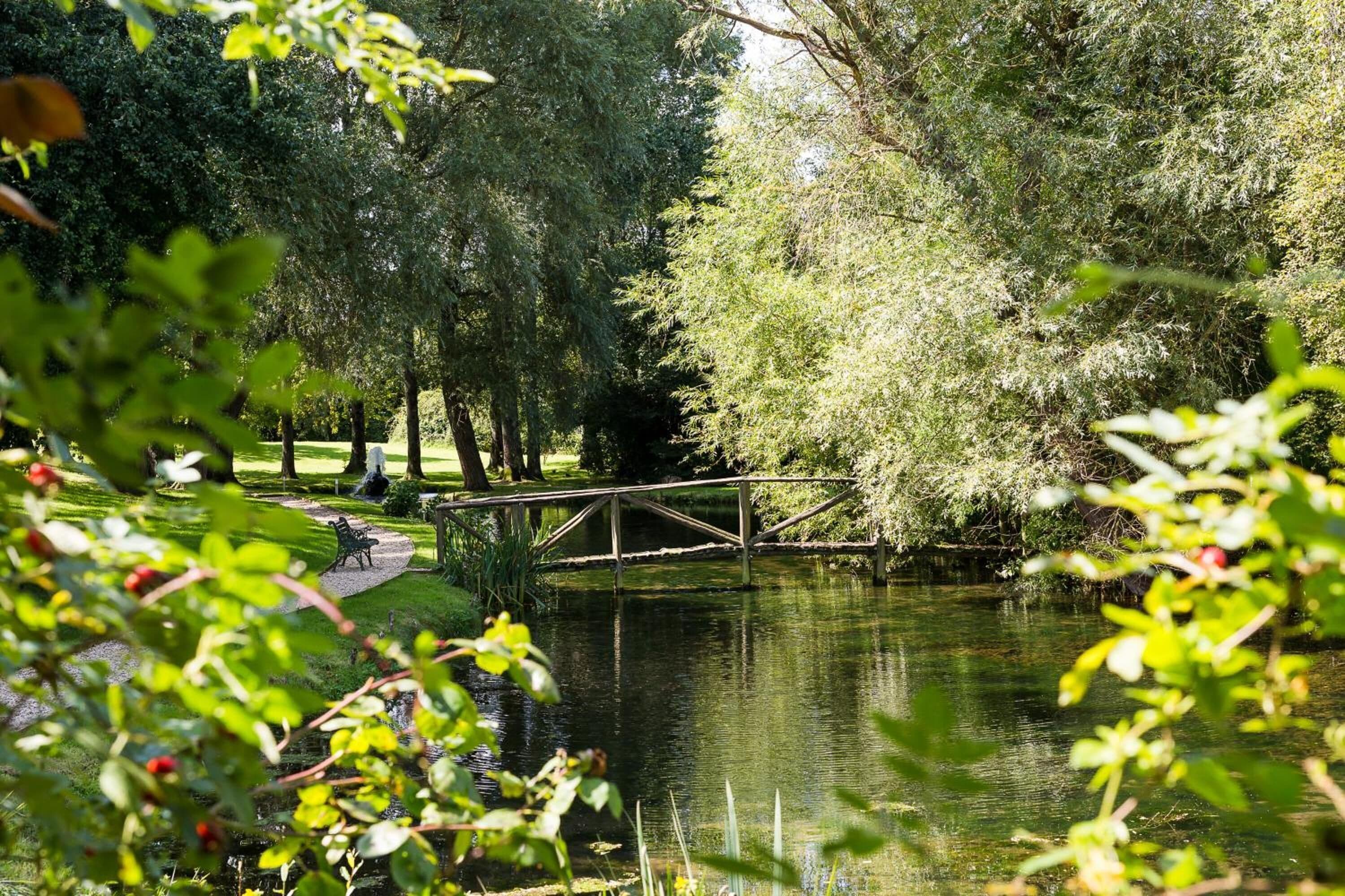 A serene park with a wooden bridge over a calm pond, surrounded by lush greenery and trees.