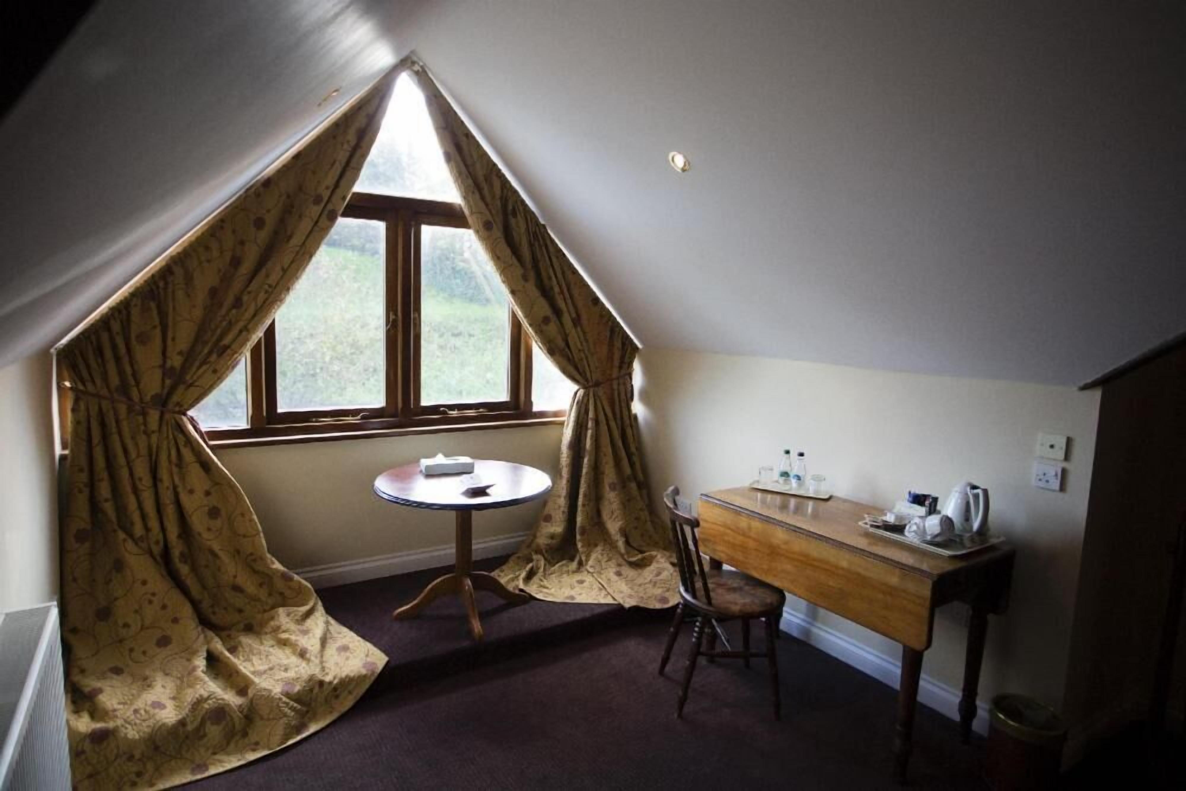 Une pièce avec un plafond en pente, une table ronde, un bureau en bois et une fenêtre avec des rideaux.