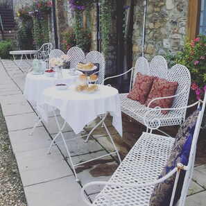 An outdoor seating area with white metal chairs and tables, covered with white tablecloths, and adorned with flowers and pastries. - Hornsbury Mill (Chard)