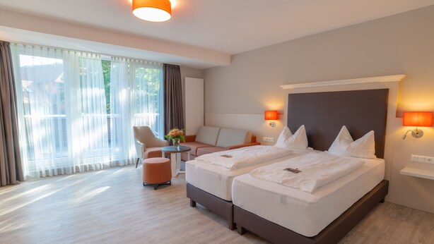 A hotel room with two beds, a sofa, a chair, a small table, and a window with curtains. - Flair Hotel Rieckmann (Bispingen)