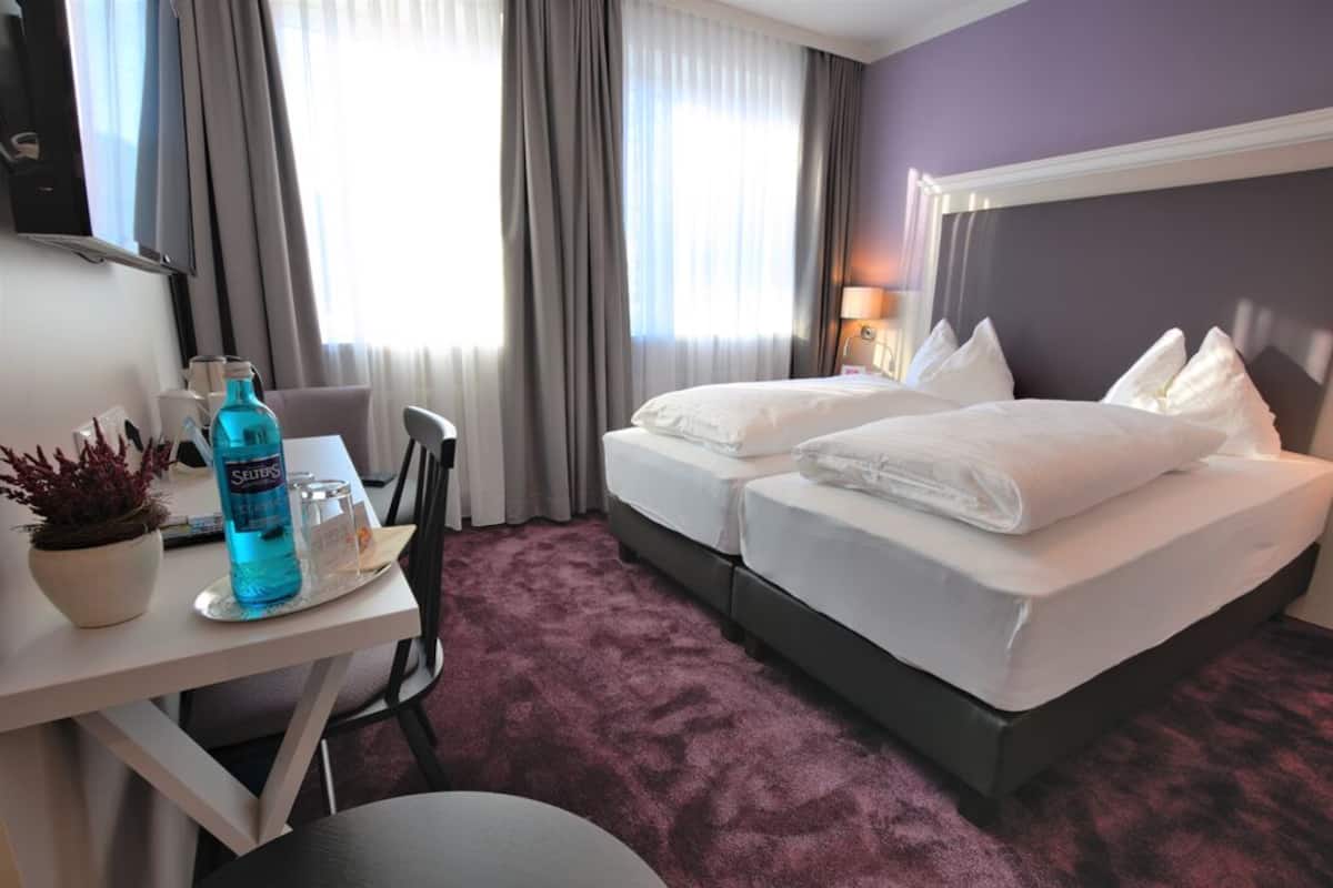 A hotel room with two beds, a desk with a bottle of water, a plant, and a TV on the wall.