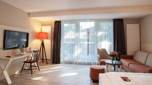 A hotel room with a flat-screen TV, a desk with a chair, a sofa, a small round table with a vase of flowers, and a bed. - Flair Hotel Rieckmann (Bispingen)