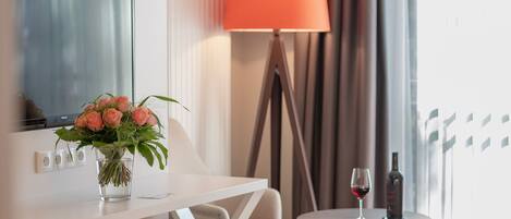 Habitación de hotel con cama, mesita de noche con un jarrón de flores, lámpara y una pequeña mesa redonda con una copa de vino y una botella.