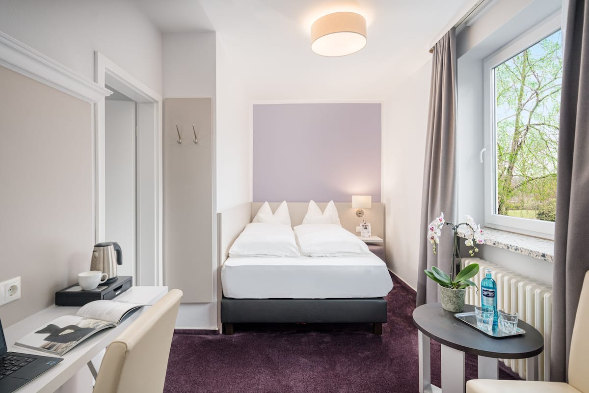 A hotel room with a bed, a desk with a laptop, a small table with a water bottle, and a window with a view of trees.