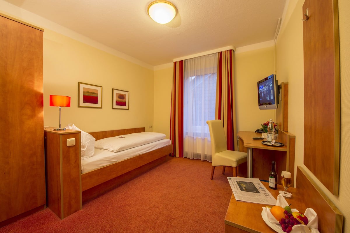 A hotel room with a bed, a desk with a computer, a chair, a lamp, and a window with curtains.