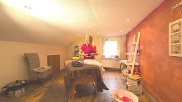 Una habitación con una camilla de masaje, una silla de mimbre, una mesita con un cuenco, un radiador y una ventana con cortinas.
