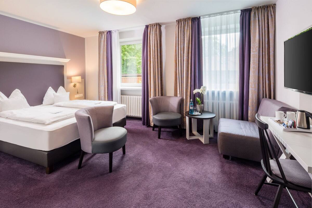 A hotel room with a bed, two armchairs, a small table with a vase of flowers, and a TV.