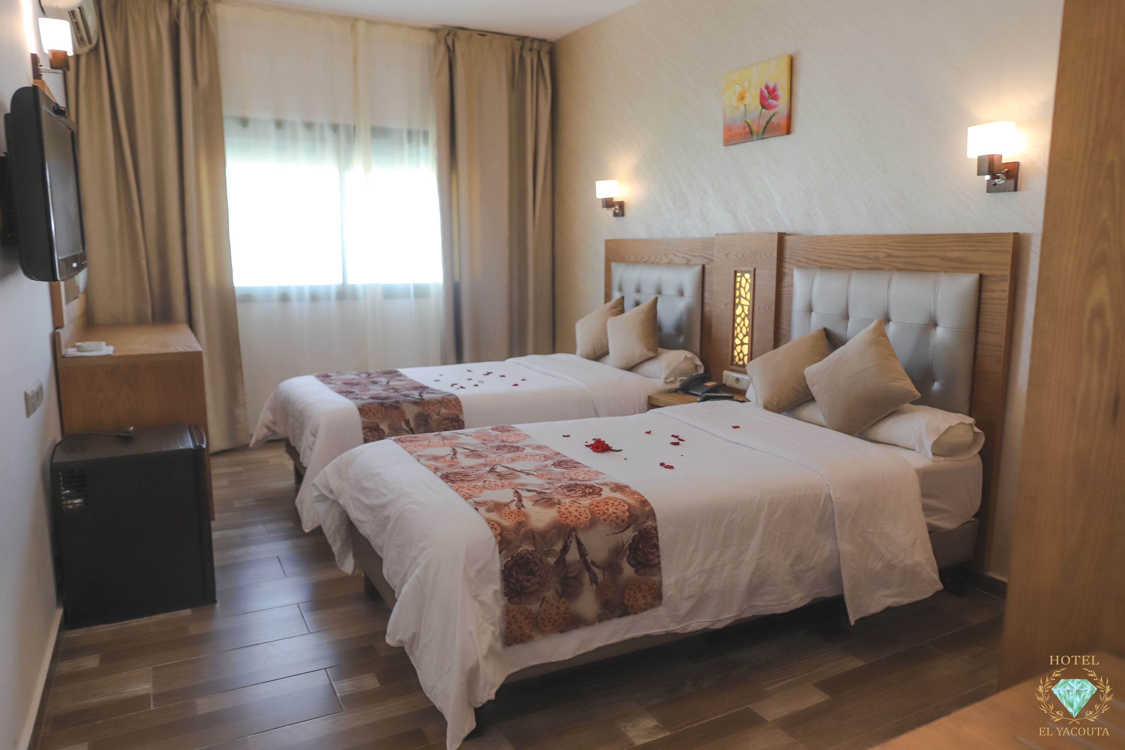 A hotel room with two beds, a TV, a small desk, and a window with curtains.