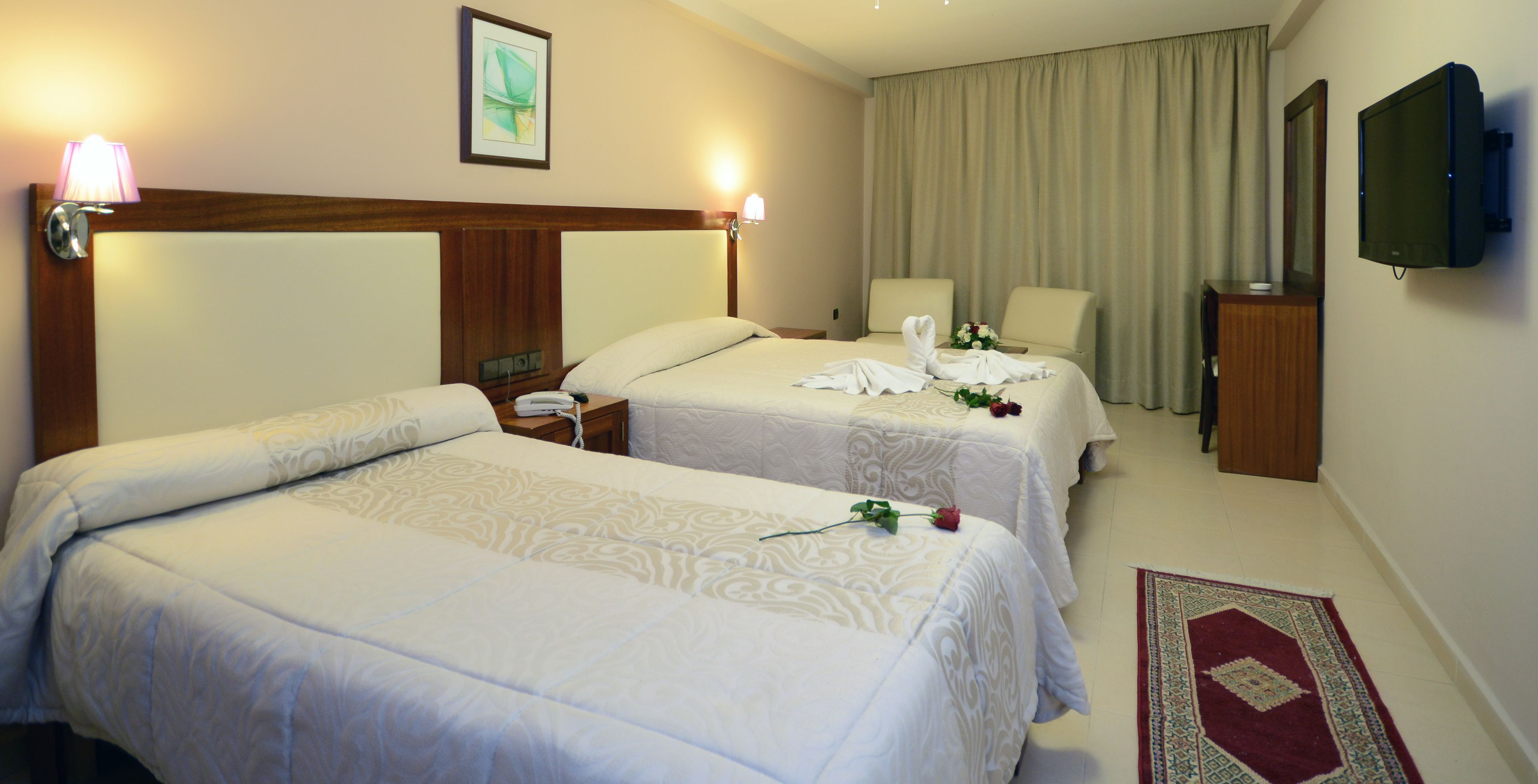 A hotel room with two beds, a TV, a bedside table with a lamp, and a red carpet.
