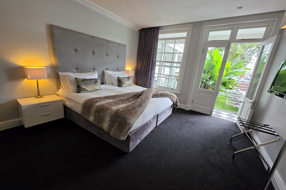 A hotel room with a large bed, a nightstand with a lamp, a view of the outdoors through a glass door, and a TV mounted on the wall.