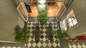 A checkered floor with two ornate tables and potted plants, a wooden ceiling, and a view into a room with a wooden floor.