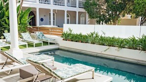 A pool area with lounge chairs and a small seating area, surrounded by greenery and a white building in the background.