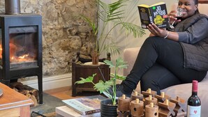 A person reading a book in a cozy room with a fireplace, a potted plant, and a wine bottle on the table.