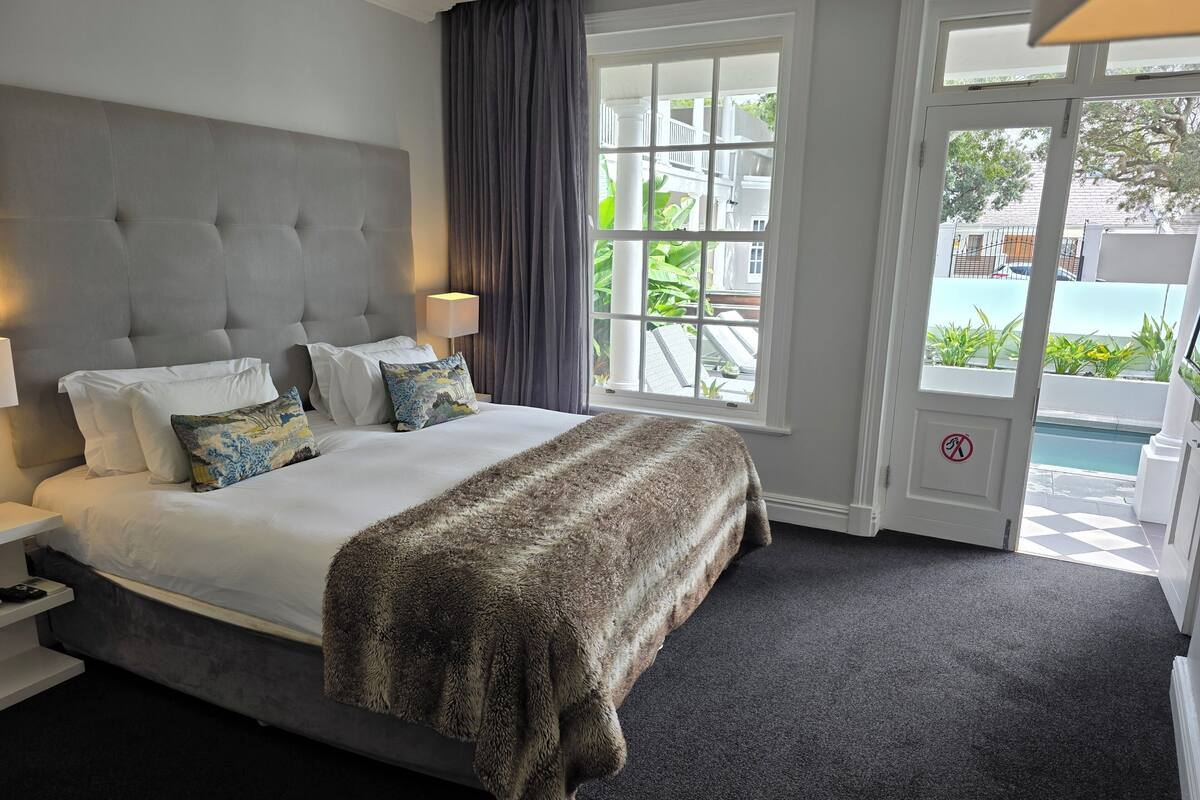 A hotel room with a large bed, a grey headboard, white bedding, and a view of a swimming pool through a glass door.