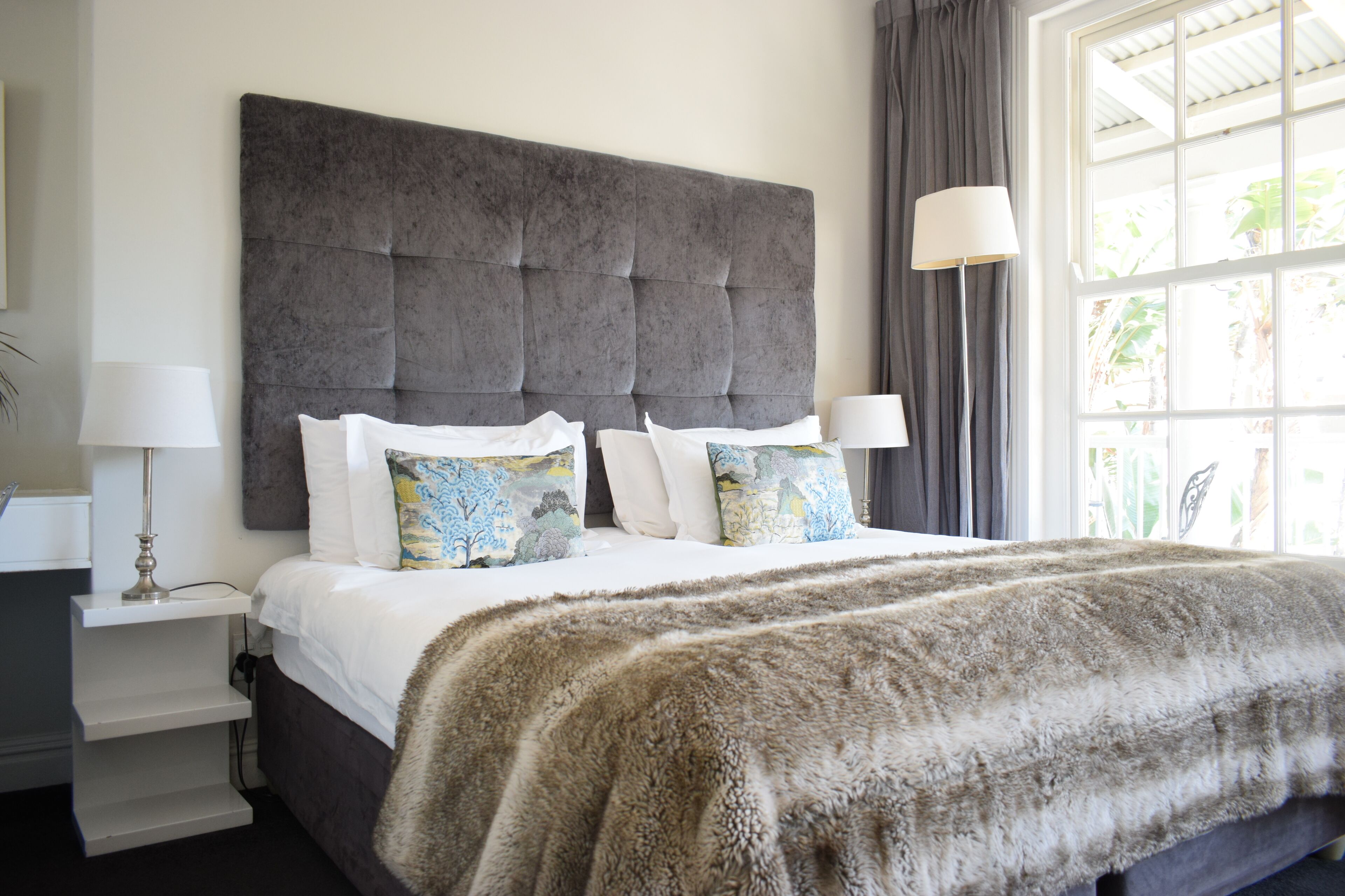 Un dormitorio con un gran cabecero capitonado, una colcha blanca y almohadones decorativos. Hay una mesita de noche con una lámpara y una lámpara de pie cerca de la ventana.