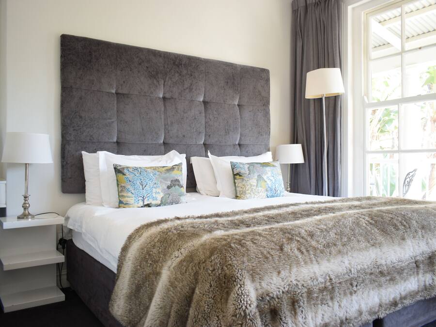 Un dormitorio con un gran cabecero capitonado, una colcha blanca y almohadones decorativos. Hay una mesita de noche con una lámpara y una lámpara de pie cerca de la ventana.
