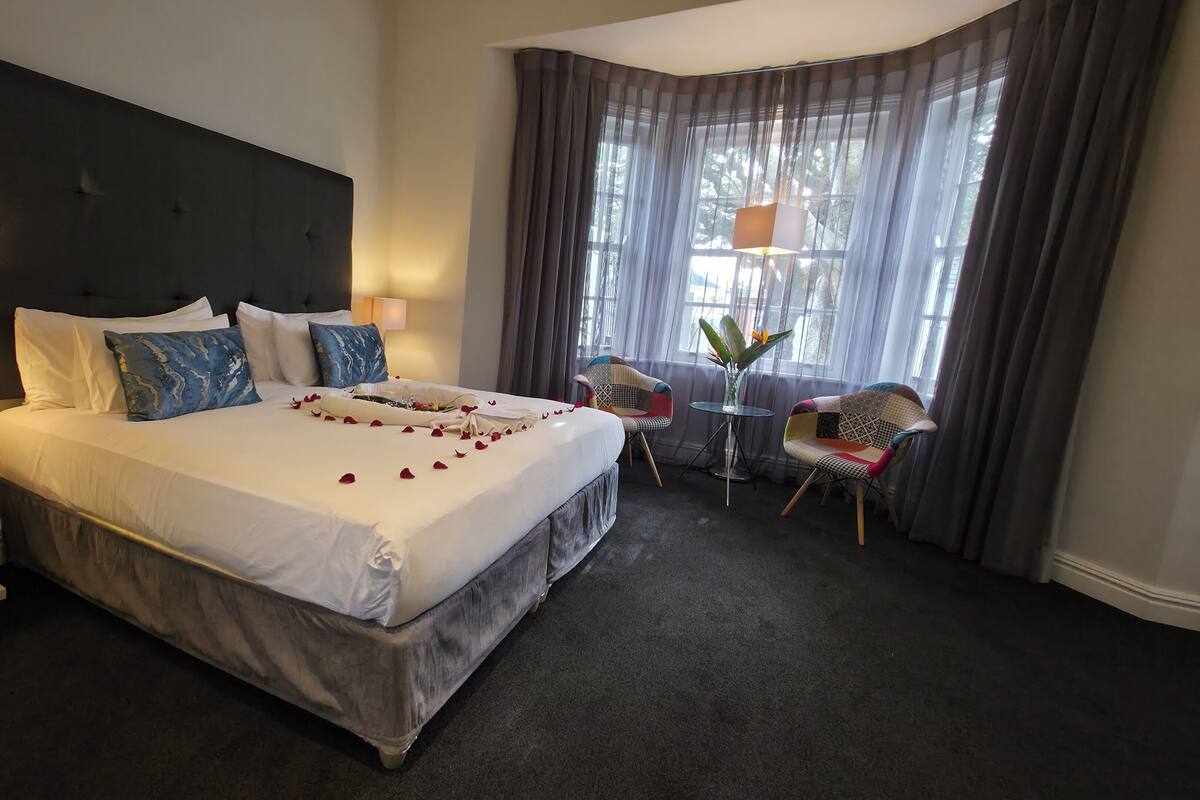 A hotel room with a large bed, a bedside table with a lamp, a small table with a vase, two chairs, and a window with curtains.