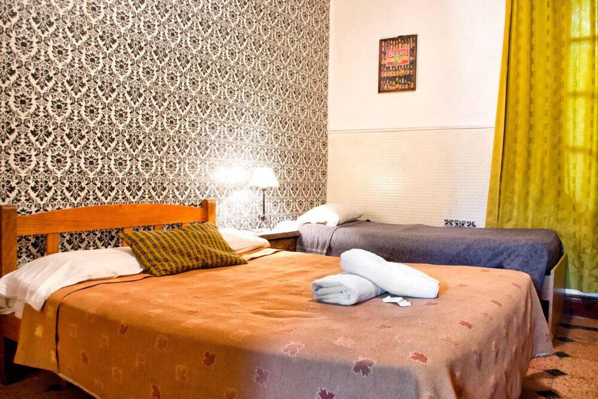 A hotel room with two beds, a patterned wall, a poster, and a yellow curtain.