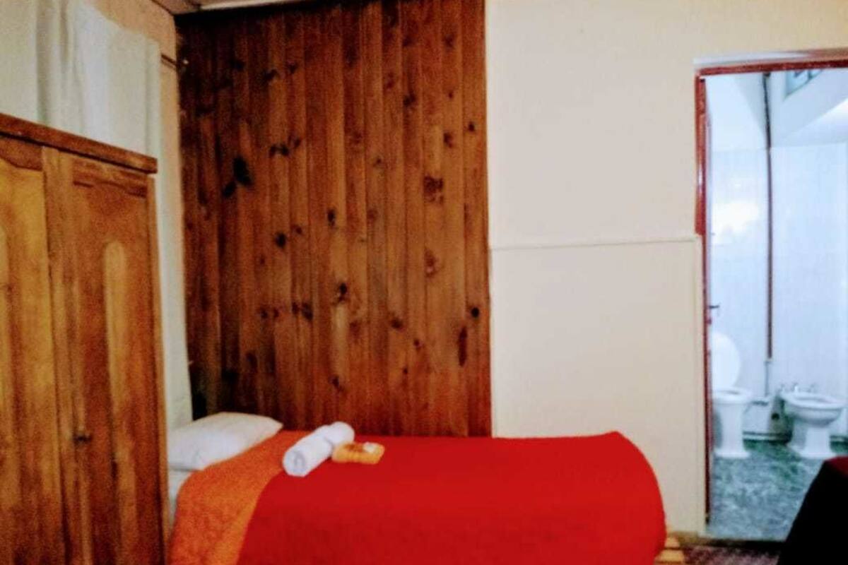 A room with a wooden wardrobe, a bed with a red blanket, and a view of toilets through a glass door.